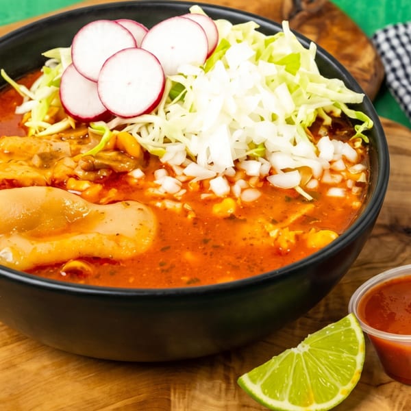 Pozole rojo in a black bowl, topped with cabbage, radishes, and onions, with a lime wedge and salsa.