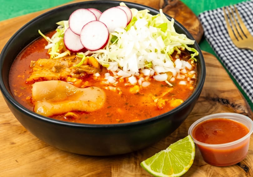 Pozole rojo in a black bowl, topped with cabbage, radishes, and onions, with a lime wedge and salsa.